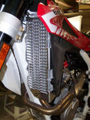A close-up of a motorcycle radiator featuring the Emperor Racing Radiator Guard. It's encased in a red and white fairing, with visible engine and exhaust pipes below. Parts of the frame and a rear-view mirror are also seen.
