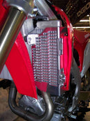 A close-up of a motorcycle radiator featuring the Emperor Racing Radiator Guard. It's encased in a red and white fairing, with visible engine and exhaust pipes below. Parts of the frame and a rear-view mirror are also seen.