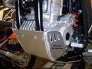 Close-up of the Emperor Racing Heavy Duty Skid Plate with a scorpion design, made from durable aluminum and installed under the engine. Its polished surface and visible bolts contrast with a blurred wooden fence background—ideal for off-road enthusiasts.