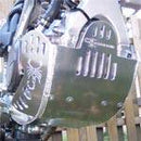Close-up of the Emperor Racing Heavy Duty Skid Plate with a scorpion design, made from durable aluminum and installed under the engine. Its polished surface and visible bolts contrast with a blurred wooden fence background—ideal for off-road enthusiasts.