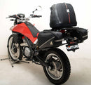 A blue adventure motorcycle, equipped with dual exhausts and off-road tires, is parked on a stand against a white background. It features a windshield and a black rear-mounted Tough-Rack by Ventura for luggage.