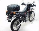A blue adventure motorcycle, equipped with dual exhausts and off-road tires, is parked on a stand against a white background. It features a windshield and a black rear-mounted Tough-Rack by Ventura for luggage.