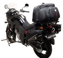 A blue adventure motorcycle, equipped with dual exhausts and off-road tires, is parked on a stand against a white background. It features a windshield and a black rear-mounted Tough-Rack by Ventura for luggage.