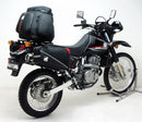 A blue adventure motorcycle, equipped with dual exhausts and off-road tires, is parked on a stand against a white background. It features a windshield and a black rear-mounted Tough-Rack by Ventura for luggage.