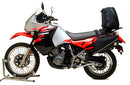 A blue adventure motorcycle, equipped with dual exhausts and off-road tires, is parked on a stand against a white background. It features a windshield and a black rear-mounted Tough-Rack by Ventura for luggage.