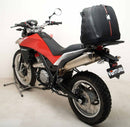 A blue adventure motorcycle, equipped with dual exhausts and off-road tires, is parked on a stand against a white background. It features a windshield and a black rear-mounted Tough-Rack by Ventura for luggage.