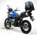 A blue adventure motorcycle, equipped with dual exhausts and off-road tires, is parked on a stand against a white background. It features a windshield and a black rear-mounted Tough-Rack by Ventura for luggage.