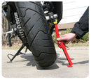 A person uses a Snapjack from Tirox to stabilize the rear tire of a motorcycle with a red wheel cleaning stand, keeping it lifted for maintenance on a concrete surface, with a partial building view in the background.