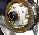 A close-up shot features an Oggy Knobbs Ducati Axle sprocket: golden teeth with silver bolts and a black hub, encircled by the chain. Parts of the wheel and tire are visible in the background.