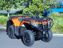 A Ventura CF Moto C-Force 500 EPS (22 - >) ATV in orange with black bull-bars is parked on a paved area near green shrubs and trees, with a blue vehicle in the background.