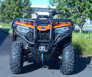 A Ventura CF Moto C-Force 500 EPS (22 - >) ATV in orange with black bull-bars is parked on a paved area near green shrubs and trees, with a blue vehicle in the background.