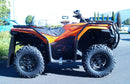 A Ventura CF Moto C-Force 500 EPS (22 - >) ATV in orange with black bull-bars is parked on a paved area near green shrubs and trees, with a blue vehicle in the background.