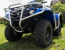 A blue Yamaha Kodiak 700 ATV, featuring a silver grille and bull bars, is parked on grass. It has large rugged tires amid trees and bushes. Under a clear sky, the vehicle by Ventura, equipped with mud flaps from the SE (16-17) series, faces slightly right.