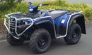 A Yamaha YFM 450 FBP Kodiak (17-21) by Ventura, in blue with a company logo and robust metal bars, is parked on pavement. It has large rugged tires and is set against lush greenery with yellow flowers.