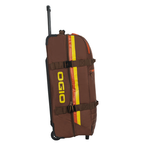 The OGIO TRUCKER Gear Bag - Stay Classy is a brown gear bag with an extended black handle, yellow and orange vertical stripes on the side, and a yellow OGIO logo. It is crafted from heavy-duty fabric, designed for carrying sports equipment, and features multiple straps and separate compartments.