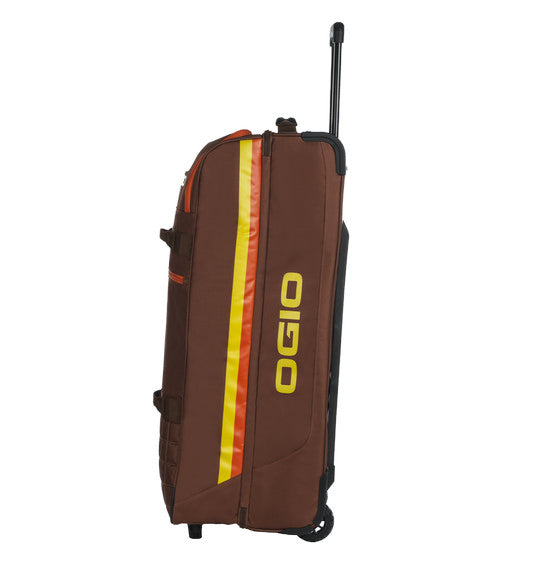 The OGIO TRUCKER Gear Bag - Stay Classy is a brown gear bag with an extended black handle, yellow and orange vertical stripes on the side, and a yellow OGIO logo. It is crafted from heavy-duty fabric, designed for carrying sports equipment, and features multiple straps and separate compartments.