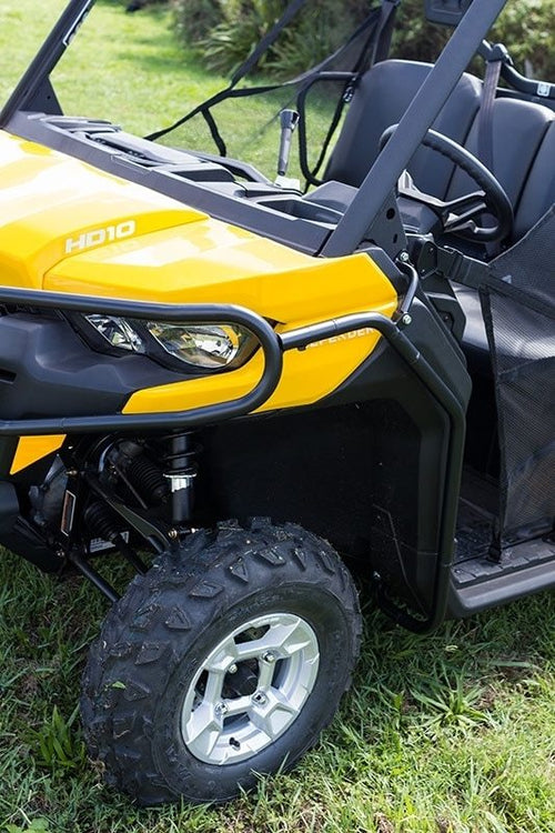 A Ventura Can-Am ATV Defender HD10 (16-17), painted yellow and black, is parked on grassy terrain. This off-road utility vehicle features rugged tires, a sturdy bull-bar, a protective front grille, mud flaps, and two side mirrors against a backdrop of greenery and a partly cloudy sky.