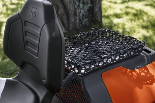 Close-up of the rear seat and cargo area of a Husqvarna all-terrain vehicle (ATV). The seat is equipped with a high backrest, and the cargo area features a sturdy bungee net for securing items. Enhanced with the Husqvarna Cargo Holder, this practical carrier boasts an innovative luggage rack kit design. In the background, tree bark and grassy area are visible.