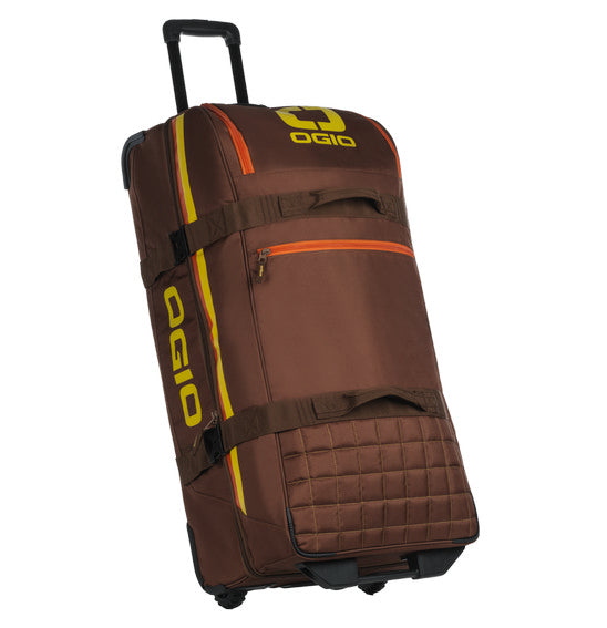 The OGIO TRUCKER Gear Bag - Stay Classy is a brown gear bag with an extended black handle, yellow and orange vertical stripes on the side, and a yellow OGIO logo. It is crafted from heavy-duty fabric, designed for carrying sports equipment, and features multiple straps and separate compartments.
