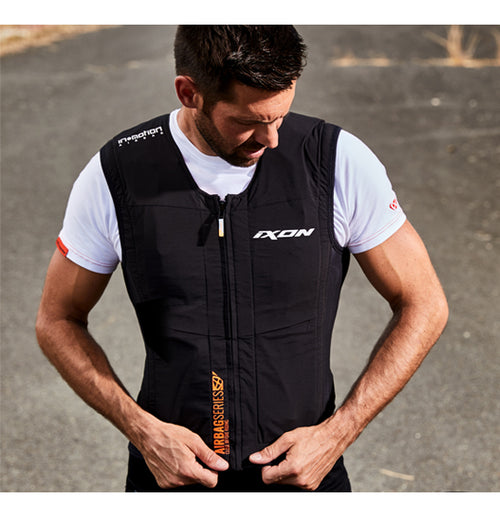 A man wearing the Ixon IX-AIRBAG U03 Airbag Vest over a white shirt adjusts the front zipper. The vest, showcasing logos such as "Ixon" and "In&motion airbag," provides advanced motorcycle protection. The background features an outdoor paved area.