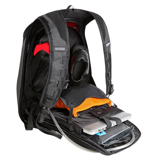 An Ogio MACH 5 Motorcycle Backpack - Stealth is open, revealing various compartments filled with items. The main compartment contains a helmet and a laptop, while smaller pockets hold a tablet and other accessories. Its interior has orange and black sections for organized storage, showcasing No Drag Technology.