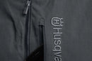 The Husqvarna Xplorer Shell Jacket Charcoal Black - Women by Husqvarna is a dark gray, hooded outdoor jacket. This windproof jacket features a full front zipper, zippered pockets, and subtle branding on the chest. It has long sleeves, adjustable ventilation, and a slightly fitted design, making it suitable for various weather conditions.