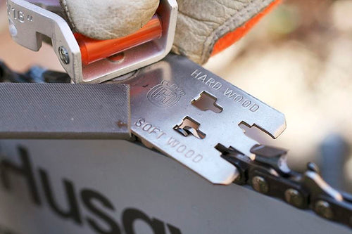 A small, blue and metallic Husqvarna Combination File Guide and Depth Gauge with cylindrical rollers. The metal piece features a flat, irregularly shaped extension with cutouts, indicating its use as a mechanical or mounting device possibly for a chain filing kit. The Husqvarna device rests on a white surface with a slight shadow underneath.