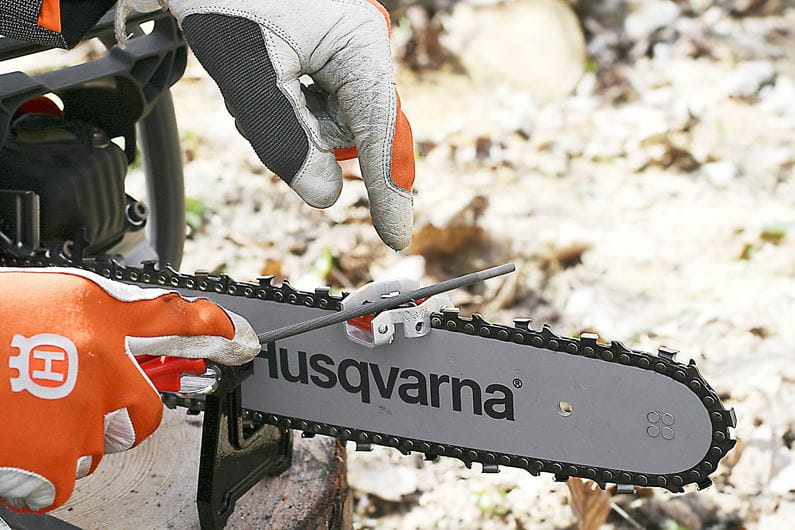 A small, blue and metallic Husqvarna Combination File Guide and Depth Gauge with cylindrical rollers. The metal piece features a flat, irregularly shaped extension with cutouts, indicating its use as a mechanical or mounting device possibly for a chain filing kit. The Husqvarna device rests on a white surface with a slight shadow underneath.