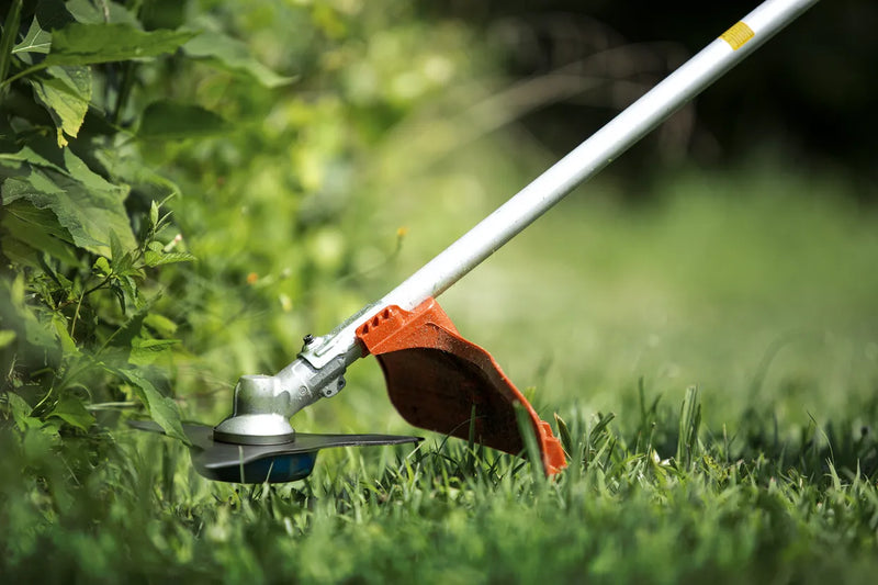 A Husqvarna 325iR Brushcutter (skin) lays on a white background. Perfect for lawns and other outdoor maintenance, the tool features a long silver shaft with a black handle at the top and an orange cutting head at the bottom. The motor is housed in an orange casing near the handle.