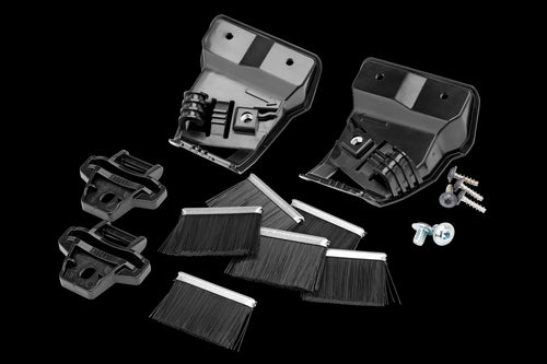 Various black and metallic car parts and accessories are laid out on a white background, including two large plastic components, smaller plastic parts, hose clamps, screws, bristle strips, and a Husqvarna Automower® Wheel Brush Kit.