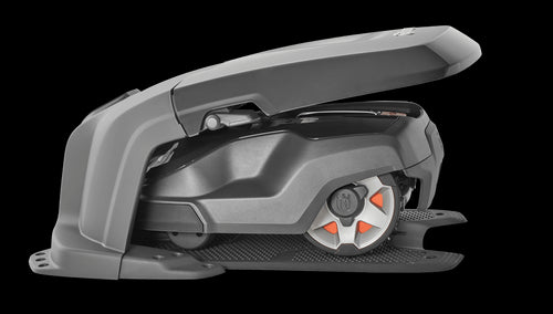 A Husqvarna AutoMower® House - 305/310/315/315X/405X/415X parked under its canopy at the edge of a lawn. The mower has a sleek, dark design with red-accented wheels. The background features an outdoor wooden deck and a row of tall, green plants, blending seamlessly with the surrounding gardens.