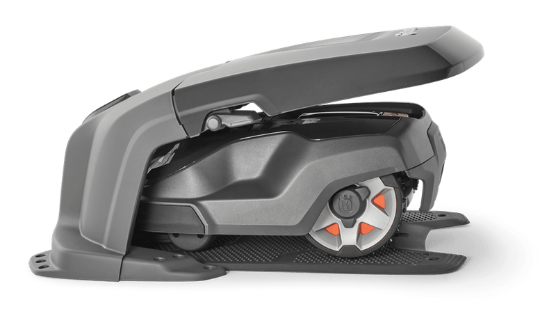 A Husqvarna AutoMower® House - 305/310/315/315X/405X/415X parked under its canopy at the edge of a lawn. The mower has a sleek, dark design with red-accented wheels. The background features an outdoor wooden deck and a row of tall, green plants, blending seamlessly with the surrounding gardens.