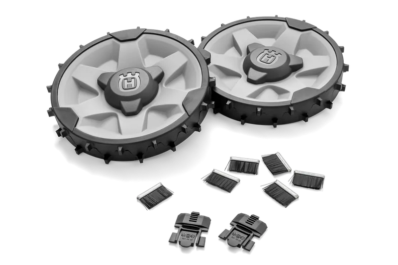 The Husqvarna Automower® Rough Terrain Kit by Husqvarna is displayed, featuring two rugged, circular robotic mower wheels with black and white treads. These treaded wheels have thick patterns suitable for rough terrain, providing excellent traction with accents in orange and gray, accompanied by a set of five small, black bristle brushes.