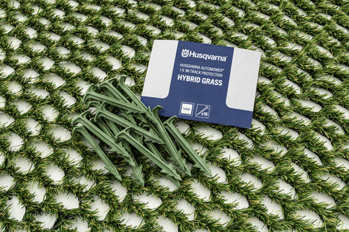 A pack of Husqvarna Automower® Hybrid Grass kit is displayed on a mesh surface. The kit includes green synthetic grass pegs and a package that reads "Husqvarna Automower® Hybrid Grass" and "11 X Extra Protection." Perfect accessories for your Husqvarna robotic lawn mower.