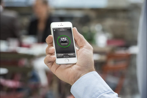 A person holds a smartphone displaying an app on its screen with an icon of a car and the word "MOVING" inside a green circle. The app, similar to the Husqvarna Automower Connect Kit by Husqvarna, has control buttons labeled "STOP" and "PARK." A blurred background shows an outdoor setting with tables and chairs.