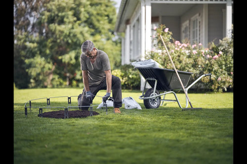 A person wearing a gray shirt and gloves is kneeling on a grassy lawn, assembling a metal frame for a small garden bed. Nearby, a Husqvarna Automower® Temporary Fence Kit and gardening materials accompany the wheelbarrow and rake. A house with a porch and flowering bushes provides the backdrop.