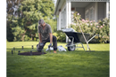 A person wearing a gray shirt and gloves is kneeling on a grassy lawn, assembling a metal frame for a small garden bed. Nearby, a Husqvarna Automower® Temporary Fence Kit and gardening materials accompany the wheelbarrow and rake. A house with a porch and flowering bushes provides the backdrop.