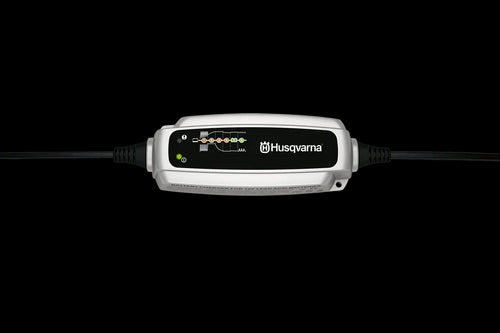 A Husqvarna Garden Tractor Battery Charger BC 0.8 against a black background. The device is rectangular, predominantly white and black with control indicators and the Husqvarna logo on the front. It has connectors on both sides for attaching to a bike or motorbike battery.