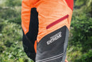 An image of Husqvarna Technical Extreme Waist trousers, high-visibility orange and black protective work pants with adjustable suspenders. These pants, made by Husqvarna, feature Cordura reinforcement at the knees and multiple pockets. The suspenders are adjustable and include reflective strips for enhanced visibility, making them ideal for forest work.
