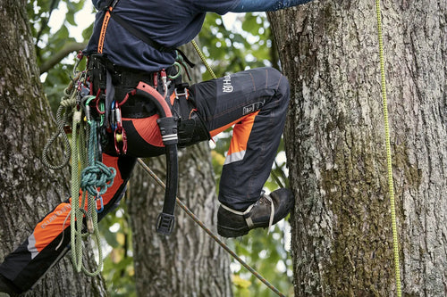 A pair of Husqvarna Arbor Waist trousers, Technical Extreme, in orange and black with attached suspenders designed for climbing and working in trees. These trousers feature multiple pockets, reinforced knee areas, and adjustable straps branded with "Husqvarna." The rugged design offers safety, agility, and functionality for demanding tasks.