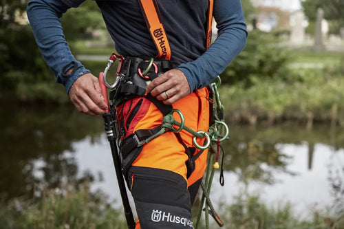 A pair of Husqvarna Arbor Waist trousers, Technical Extreme, in orange and black with attached suspenders designed for climbing and working in trees. These trousers feature multiple pockets, reinforced knee areas, and adjustable straps branded with "Husqvarna." The rugged design offers safety, agility, and functionality for demanding tasks.