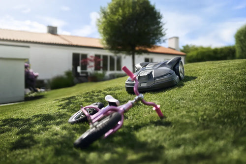 The Husqvarna Automower 430X NERA is a sleek, grey robotic lawn mower featuring two large wheels at the rear and two smaller wheels at the front. It boasts a red stop button on top, LED headlights, and proudly displays the Husqvarna logo on the front. With its modern design, this mower is equipped with object avoidance technology and can be conveniently controlled via the Automower Connect app.