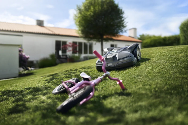 The Husqvarna Automower 430X NERA is a sleek, grey robotic lawn mower featuring two large wheels at the rear and two smaller wheels at the front. It boasts a red stop button on top, LED headlights, and proudly displays the Husqvarna logo on the front. With its modern design, this mower is equipped with object avoidance technology and can be conveniently controlled via the Automower Connect app.