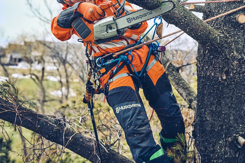 The Husqvarna T542i XP Battery Chainsaw (Skin Only) features an eye-catching orange and black body, a sturdy metal chain bar with the "Husqvarna" brand name displayed on it, a grip handle, and various adjustment controls against a transparent background.
