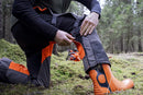 The Husqvarna Functional Safety Chaps, designed in bright orange and black for enhanced safety, feature durable fabric with reinforced areas. The high-visibility "Husqvarna" brand name is prominently displayed on the left thigh. These chaps offer an adjustable fit and boast a modern, rugged look ideal for outdoor work.