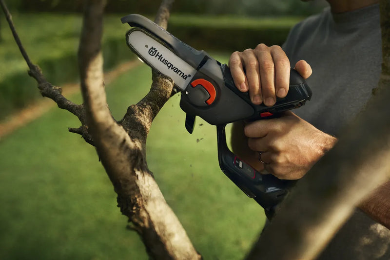 A black and orange, battery-powered telescopic pole saw with a small chainsaw attachment on the end, ideal for outdoor trimming tasks. The set includes a durable Husqvarna battery pack and a separate battery charger, all neatly arranged on a plain, white background. Perfect for maintaining lawns and gardens.