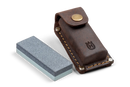 A Husqvarna Axe Grinding Stone, made of rectangular silicon carbide with a dual-color surface of grey and white, is placed next to a brown leather sheath featuring a snap button and stitched edges. The sheath, adorned with an embossed Husqvarna logo, stands out against the completely black background.