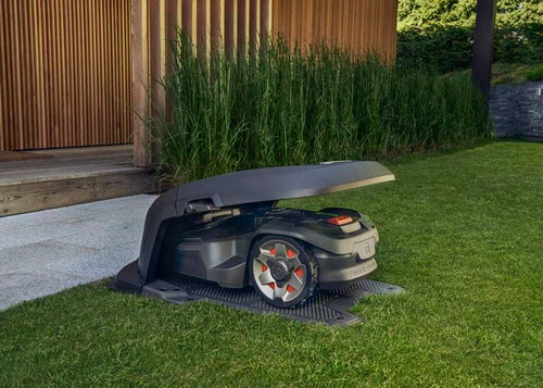 A Husqvarna AutoMower® House - 305/310/315/315X/405X/415X parked under its canopy at the edge of a lawn. The mower has a sleek, dark design with red-accented wheels. The background features an outdoor wooden deck and a row of tall, green plants, blending seamlessly with the surrounding gardens.