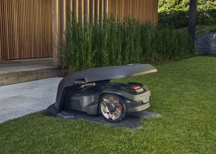 A Husqvarna AutoMower® House - 305/310/315/315X/405X/415X parked under its canopy at the edge of a lawn. The mower has a sleek, dark design with red-accented wheels. The background features an outdoor wooden deck and a row of tall, green plants, blending seamlessly with the surrounding gardens.