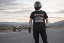Person wearing a black t-shirt with a motorcycle graphic and text, standing on a road with another motorcycle in the background.